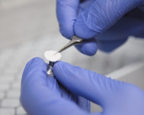 Gloved hands carefully handle a small, white sample with tweezers. Photo Source: University of New Mexico
