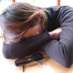 Representative Image: A person sitting at a table with their head resting on their folded arms, suggesting they might be tired, sleeping, or feeling overwhelmed. Photo Source: Les Rififis (CC BY-NC-ND 2.0)