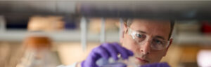 A scientist or researcher wearing protective glasses and purple gloves, carefully handling a sample or conducting an experiment in a laboratory setting. Photo Source: Lilly Investors.