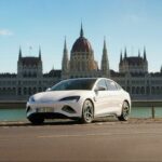 A white BYD Seal electric sedan parked beside the Danube River in front of the Hungarian Parliament Building in Budapest.