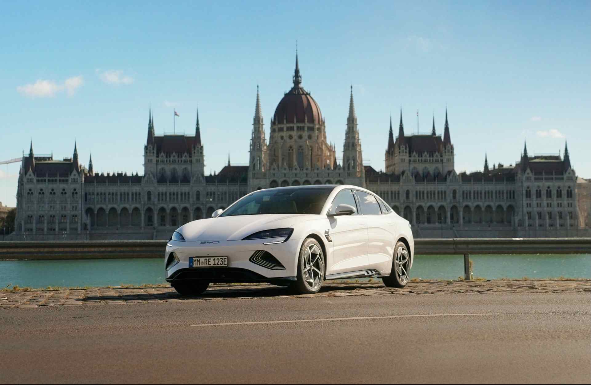 A white BYD Seal electric sedan parked beside the Danube River in front of the Hungarian Parliament Building in Budapest.