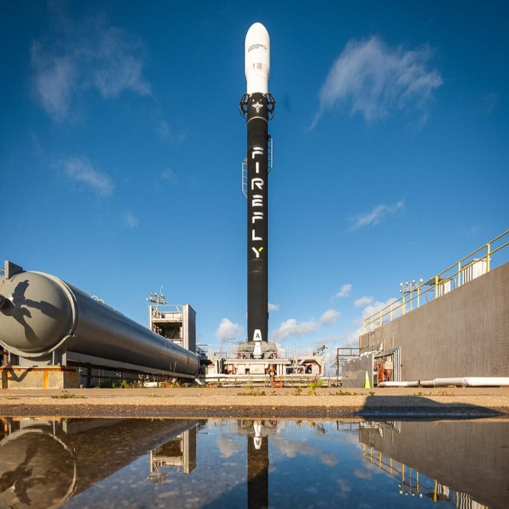 A Firefly Aerospace Alpha rocket standing vertical on the launch pad at Vandenberg Space Force Base, reflected in a water puddle beneath it.