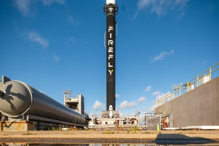 A Firefly Aerospace Alpha rocket standing vertical on the launch pad at Vandenberg Space Force Base, reflected in a water puddle beneath it.