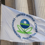 EPA flag featuring the agency’s seal—a stylized two-leaved flower beneath a sphere symbolizing sky, water, and earth—flying outside a government building. Photo Source: Paul A. Fagan (CC BY 2.0)