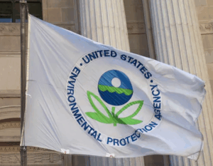 EPA flag featuring the agency’s seal—a stylized two-leaved flower beneath a sphere symbolizing sky, water, and earth—flying outside a government building. Photo Source: Paul A. Fagan (CC BY 2.0)