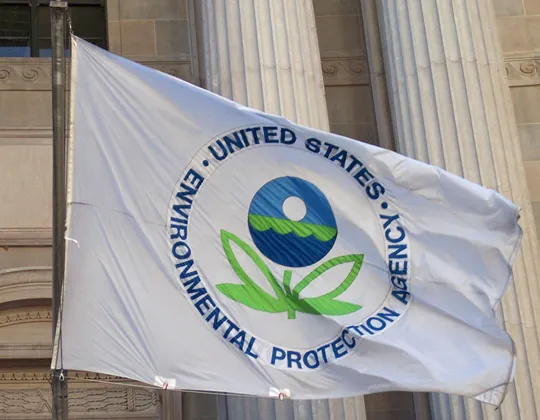 EPA flag featuring the agency’s seal—a stylized two-leaved flower beneath a sphere symbolizing sky, water, and earth—flying outside a government building. Photo Source: Paul A. Fagan (CC BY 2.0)