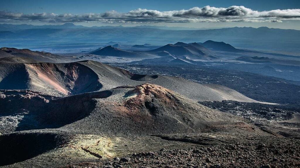 Panoramic view of barren volcanic cones, craters, and lava fields on Russia’s Kamchatka Peninsula beneath a cloudy sky, illustrating the region’s active tectonic setting.