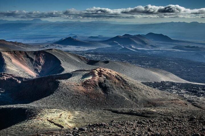 Panoramic view of barren volcanic cones, craters, and lava fields on Russia’s Kamchatka Peninsula beneath a cloudy sky, illustrating the region’s active tectonic setting.