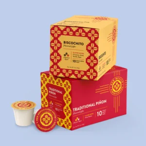 Two stacked boxes of New Mexico Piñon Coffee single‑serve pods—Biscochito flavor on top and Traditional Piñon below—alongside one exposed pod, all against a pale blue background.