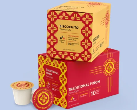Two stacked boxes of New Mexico Piñon Coffee single‑serve pods—Biscochito flavor on top and Traditional Piñon below—alongside one exposed pod, all against a pale blue background.