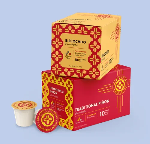 Two stacked boxes of New Mexico Piñon Coffee single‑serve pods—Biscochito flavor on top and Traditional Piñon below—alongside one exposed pod, all against a pale blue background.