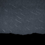 Silhouette of distant hills beneath a dark sky studded with dozens of faint, parallel meteor streaks captured in a single composite exposure.