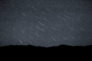 Silhouette of distant hills beneath a dark sky studded with dozens of faint, parallel meteor streaks captured in a single composite exposure.