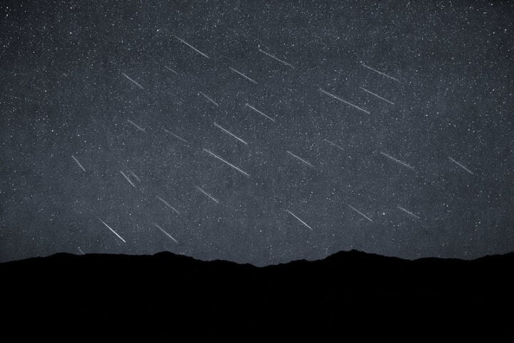 Silhouette of distant hills beneath a dark sky studded with dozens of faint, parallel meteor streaks captured in a single composite exposure.