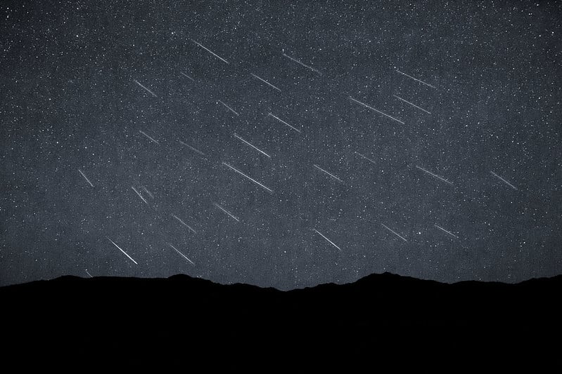 Silhouette of distant hills beneath a dark sky studded with dozens of faint, parallel meteor streaks captured in a single composite exposure.