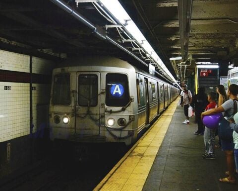 Subway platform at West 4th Street station with an A train stopped and passengers waiting under fluorescent lights.