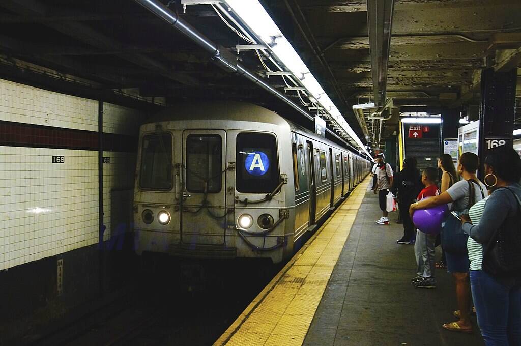 Subway platform at West 4th Street station with an A train stopped and passengers waiting under fluorescent lights.