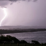 A bright lightning bolt strikes the ocean off a darkened NSW shoreline under a stormy sky.