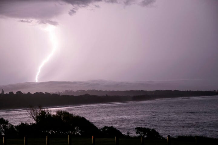 A bright lightning bolt strikes the ocean off a darkened NSW shoreline under a stormy sky.
