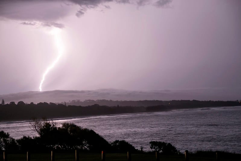 A bright lightning bolt strikes the ocean off a darkened NSW shoreline under a stormy sky.