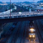 A Norfolk Southern locomotive moves beneath a highway overpass at dusk in Atlanta’s Inman Yard, surrounded by rows of intermodal containers.