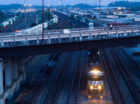 A Norfolk Southern locomotive moves beneath a highway overpass at dusk in Atlanta’s Inman Yard, surrounded by rows of intermodal containers.