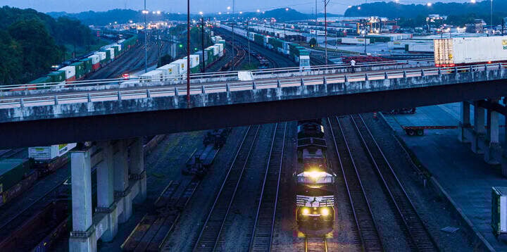 A Norfolk Southern locomotive moves beneath a highway overpass at dusk in Atlanta’s Inman Yard, surrounded by rows of intermodal containers.