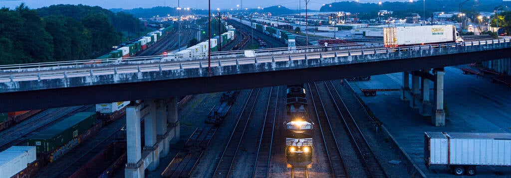 A Norfolk Southern locomotive moves beneath a highway overpass at dusk in Atlanta’s Inman Yard, surrounded by rows of intermodal containers.