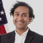 Portrait of Dr. Vinay Prasad smiling in a dark suit and white shirt, standing before an American flag.