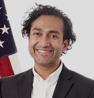 Portrait of Dr. Vinay Prasad smiling in a dark suit and white shirt, standing before an American flag.