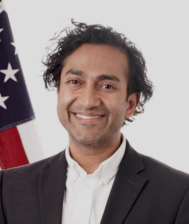 Portrait of Dr. Vinay Prasad smiling in a dark suit and white shirt, standing before an American flag.