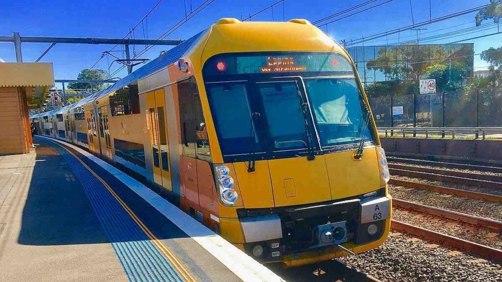 A yellow-and-steel Sydney Trains Waratah A-set (A63) standing at a suburban platform under overhead wires and clear blue sky. Photo Source: Trainsandtech (CC BY-SA 4.0)