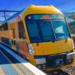 A yellow-and-steel Sydney Trains Waratah A-set (A63) standing at a suburban platform under overhead wires and clear blue sky. Photo Source: Trainsandtech (CC BY-SA 4.0)