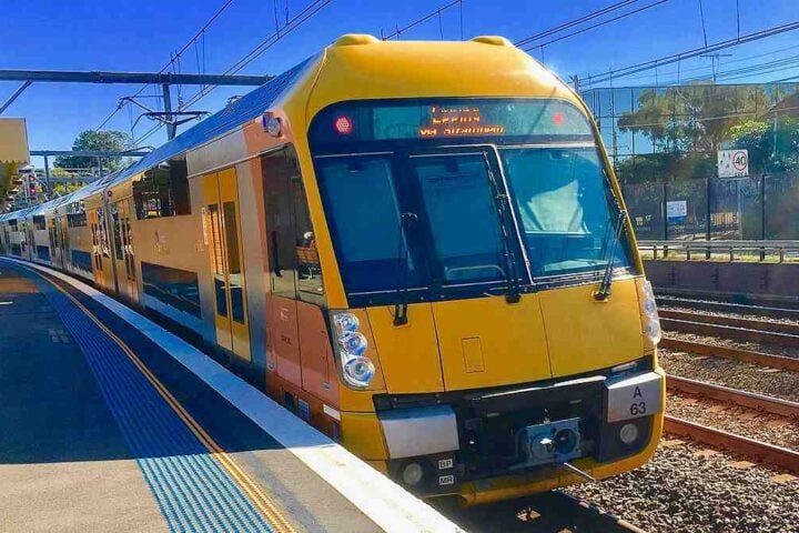 A yellow-and-steel Sydney Trains Waratah A-set (A63) standing at a suburban platform under overhead wires and clear blue sky. Photo Source: Trainsandtech (CC BY-SA 4.0)