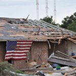 American flag draped over a heavily damaged brick home with scattered debris after a tornado.