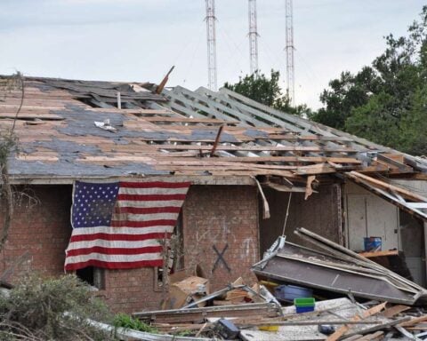 American flag draped over a heavily damaged brick home with scattered debris after a tornado.