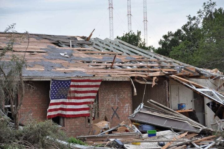 American flag draped over a heavily damaged brick home with scattered debris after a tornado.