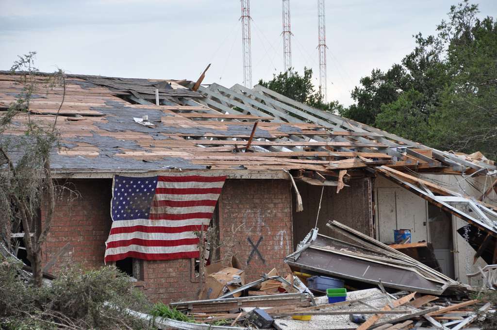 American flag draped over a heavily damaged brick home with scattered debris after a tornado.