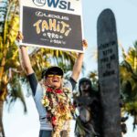 Smiling young surfer Kelia Gallina holds aloft a “WSL Lexus Tahiti Pro Wild Card Femme” sign, adorned with celebratory flower lei.