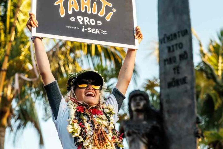 Smiling young surfer Kelia Gallina holds aloft a “WSL Lexus Tahiti Pro Wild Card Femme” sign, adorned with celebratory flower lei.