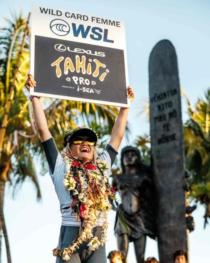 Smiling young surfer Kelia Gallina holds aloft a “WSL Lexus Tahiti Pro Wild Card Femme” sign, adorned with celebratory flower lei.