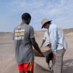 Two researchers from the Ledi-Geraru Research Project carrying a wooden box across an arid landscape in Ethiopia's Afar Region, with one wearing a gray t-shirt displaying "LEDI-GERARU RESEARCH PROJECT" on the back.