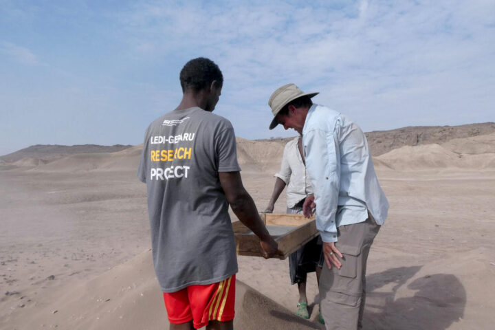 Two researchers from the Ledi-Geraru Research Project carrying a wooden box across an arid landscape in Ethiopia's Afar Region, with one wearing a gray t-shirt displaying "LEDI-GERARU RESEARCH PROJECT" on the back.