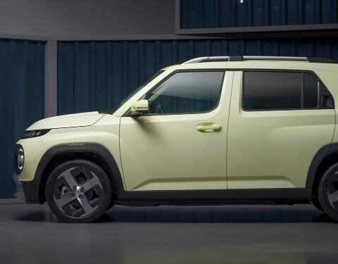 Side profile of a light-green Hyundai Inster EV parked indoors against a corrugated metal-panel backdrop.