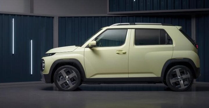 Side profile of a light-green Hyundai Inster EV parked indoors against a corrugated metal-panel backdrop.