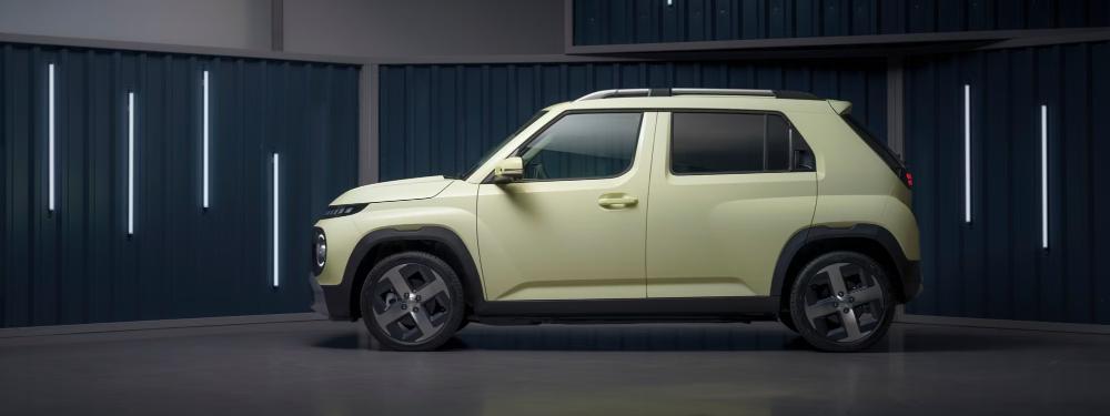 Side profile of a light-green Hyundai Inster EV parked indoors against a corrugated metal-panel backdrop.