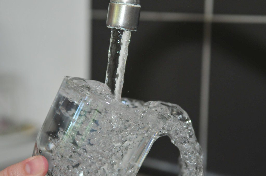 A close-up view of tap water flowing from a faucet into a glass being held by a person's hand.