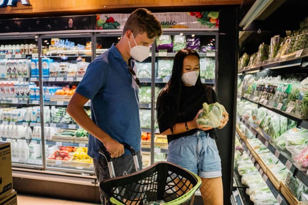 Young couple wearing face masks shopping for vegetables in a grocery store, examining lettuce while standing in front of refrigerated produce section with a shopping basket.