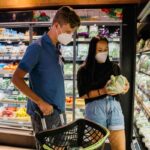 Young couple wearing face masks shopping for vegetables in a grocery store, examining lettuce while standing in front of refrigerated produce section with a shopping basket.