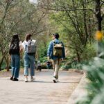 Three friends walking along a tree-lined park pathway, each carrying a backpack.